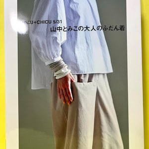 Puede incluir: Una portada de libro con una blusa azul claro y pantalones beige. El texto "NICU+CHICU 5/31" y texto japonés son visibles. El libro se titula "山中とみこの大人のふだん着" e incluye el texto "フリーサイズの縫い代つき実物大パターン".
