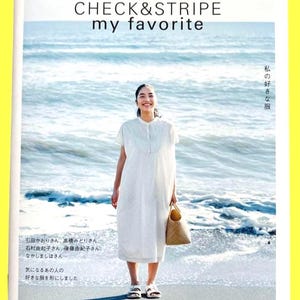 Może przedstawiać: Okładka magazynu z tekstem "CHECK & STRIPE my favorite" i japońskim tekstem. Okładka przedstawia osobę w białej sukience trzymającą plecioną torbę, stojącą na plaży z oceanem w tle.