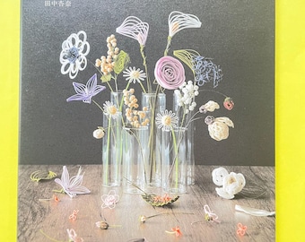 May include: A book cover featuring a dark gray background with the Japanese text 飾る・贈る・装う 花を結ぶ水引細工 and 田中杏奈. The cover displays various colorful mizuhiki cord flowers arranged in clear glass vases on a wooden surface, with additional small floral pieces scattered around the base.