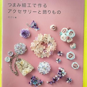 May include: A pink book cover with Japanese text and various fabric flower accessories. The accessories include a butterfly, roses, and other floral designs in shades of purple, white, and pink. The book title is "Accessories and Decorations Made with Tsumami Craft."