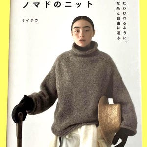 以下が含まれることがあります： グレーのタートルネックセーターを着た女性が表紙の書籍。書籍のタイトルは日本語で、追加のテキストも日本語です。女性は麦わら帽子を持ち、黒い手袋をしています。