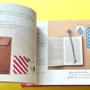 Puede incluir: Un libro abierto con un marcap&aacute;ginas azul, un marcap&aacute;ginas de lunares y un marcap&aacute;ginas decorativo rojo. El libro est&aacute; sobre una superficie beige con texto e ilustraciones japonesas. Tambi&eacute;n se muestran sobres de papel marr&oacute;n con cierres de cuerda.