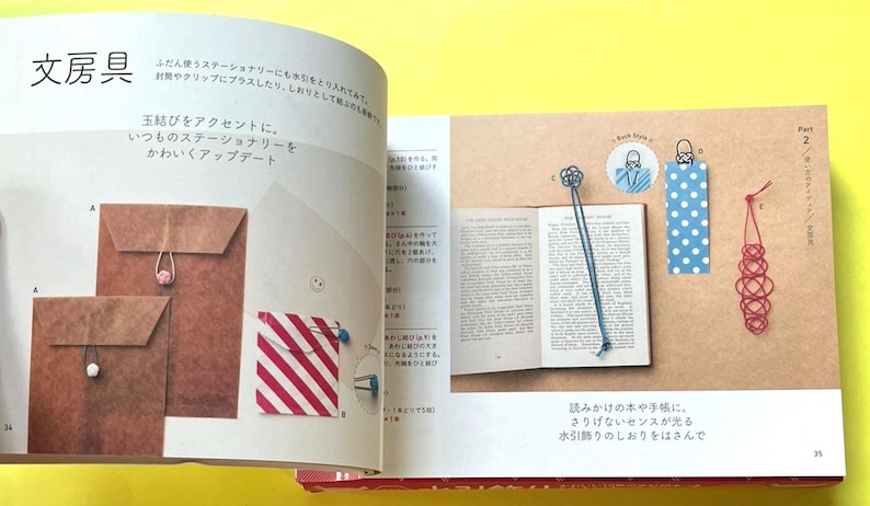 Puede incluir: Un libro abierto con un marcap&aacute;ginas azul, un marcap&aacute;ginas de lunares y un marcap&aacute;ginas decorativo rojo. El libro est&aacute; sobre una superficie beige con texto e ilustraciones japonesas. Tambi&eacute;n se muestran sobres de papel marr&oacute;n con cierres de cuerda.