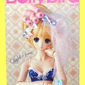 May include: A doll with blonde hair styled with pink curlers and blue bows, wearing a blue and white patterned top and a veil. The magazine cover reads "Dollybird vol.21" in pink lettering. The text "One of a kind" is also visible.