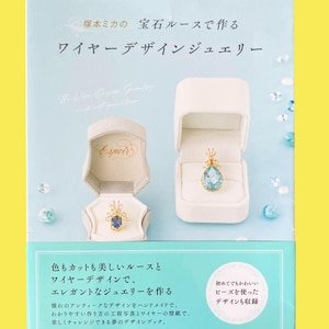 Op de afbeelding: Een boekomslag met Japanse tekst en afbeeldingen van sieraden. Twee open sieradendoosjes tonen een blauwe edelsteen hanger en een blauwe edelsteen ring. De titel van het boek is in het Japans, met extra Japanse tekst.