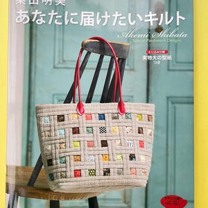 May include: A craft book titled Akemi Shibata Special Patchwork Designs featuring a woven-style patchwork tote bag with beige fabric strips and colorful patterned squares. The bag has two red leather handles and is displayed on a wooden chair.