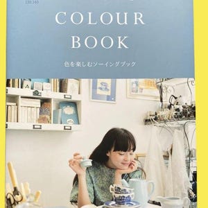May include: A book titled "COLOUR BOOK" by CHECK&STRIPE, with Japanese text. The cover is light blue. A woman is seated at a table with a tea set. The tea set includes cups, saucers, a teapot, and a creamer. The woman is holding a small, light blue object.