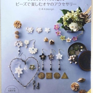 May include: A book cover in Japanese with the title "Beads for Enjoying Oya Accessories". The image displays handmade lace jewellery, including earrings and necklaces, in white, black, and gold. There are also decorative elements and a floral arrangement. Measurements are in cm.
