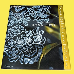 Op de afbeelding: Een boek getiteld "Cross Stitch Lace Pattern" met een zwarte kaft met een wit kantontwerp. Het boek heeft tekst in het Japans en Engels, met een gele rug. De kaft heeft ook het "applemints" logo.