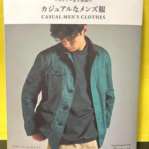 Peut inclure: Couverture de livre avec un homme portant une veste verte et une chemise sombre. Le texte sur la couverture indique "CASUAL MEN'S CLOTHES" en anglais et en japonais. Le livre fait partie de la série Heart Warming Life.
