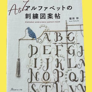 以下が含まれることがあります： 「A to Z アルファベットの刺繍図案帖」というタイトルの本の表紙。日本語と英語で書かれています。刺繍された文字、青い鳥、房が特徴です。著者の名前も表示されています。