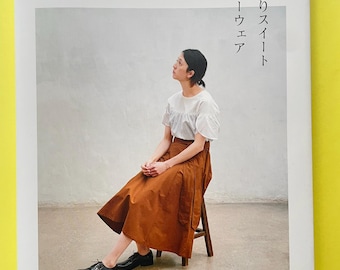 Può includere: Un libro con copertina flessibile intitolato ほんのりスイートデイリーウェア con il testo Quoi? Quoi? e S, M, L, 2L, 3L. La copertina presenta una fotografia di una persona che indossa una camicetta bianca a maniche corte e una lunga gonna a pieghe color ruggine, seduta su uno sgabello di legno.