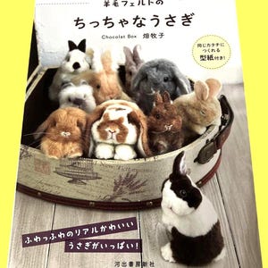 以下が含まれることがあります： 様々な色のフェルトウサギのフィギュアが飾られた、装飾的なバスケットの表紙。本のタイトルは日本語で、追加のテキストと出版社のロゴがあります。ウサギは茶色、白、灰色です。