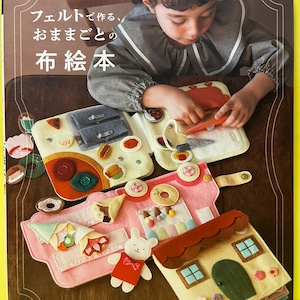 以下が含まれることがあります： フェルトで作られた布絵本で遊ぶ子供。本には、フェルトで作られた食べ物、家、その他のインタラクティブな要素が含まれています。表紙には日本語のテキストとブランド名「applemints」が表示されています。本は木製の表面に開かれています。