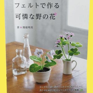 May include: A book titled "Making Cute Wildflowers with Felt" in Japanese, with a small white ceramic pot with felt flowers and a small white pitcher with felt flowers. The book is on a wooden table with two clear glass bottles.