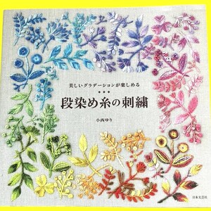 Puede incluir: Portada de libro con diseños florales bordados en degradado arcoíris. El título, en japonés, se traduce como "Bordado de hilo teñido". La portada también incluye texto japonés y el nombre del autor. El fondo es de tela beige claro.