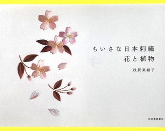 May include: A book cover with a white background and a yellow border. The cover features embroidered pink cherry blossoms and brown leaves. Japanese text is visible, along with the author's name.