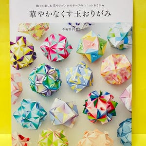 Op de afbeelding: Een softcover boek getiteld "華やかなくす玉おりがみ" met een verzameling kleurrijke, geometrische kusudama origami bollen op de kaft. Het boek toont diverse ingewikkelde papieren vouwontwerpen in tinten roze, blauw, geel en groen.