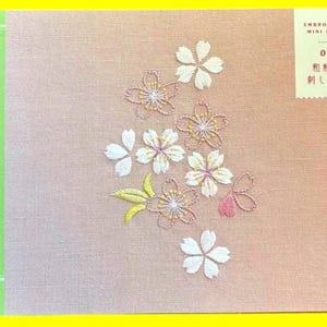 May include: A book titled "Japanese Patterns" with a green spine and a pink cover. The cover features embroidered cherry blossom designs in white, pink, and yellow. The book also has text in Japanese.