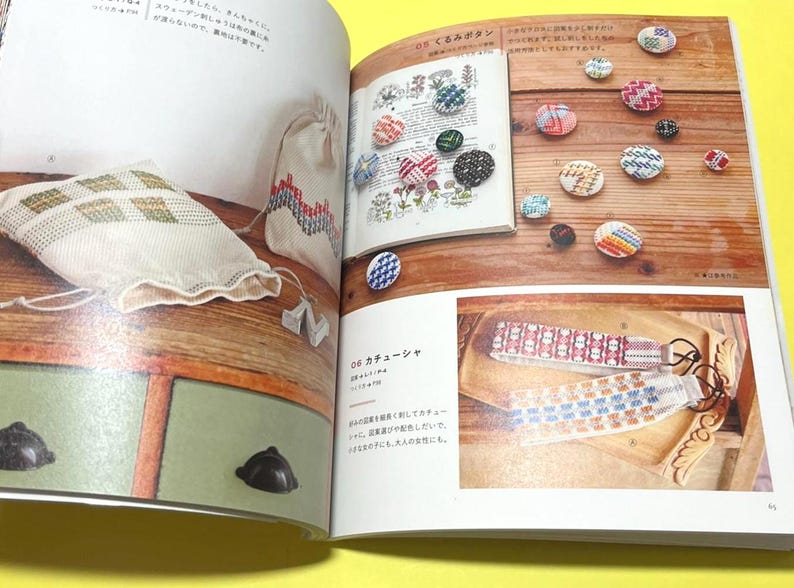 Puede incluir: Un libro abierto muestra botones hechos a mano y accesorios bordados. Una bolsa blanca y una bolsa peque&ntilde;a con patrones geom&eacute;tricos est&aacute;n sobre una superficie de madera. La p&aacute;gina tambi&eacute;n muestra diademas.
