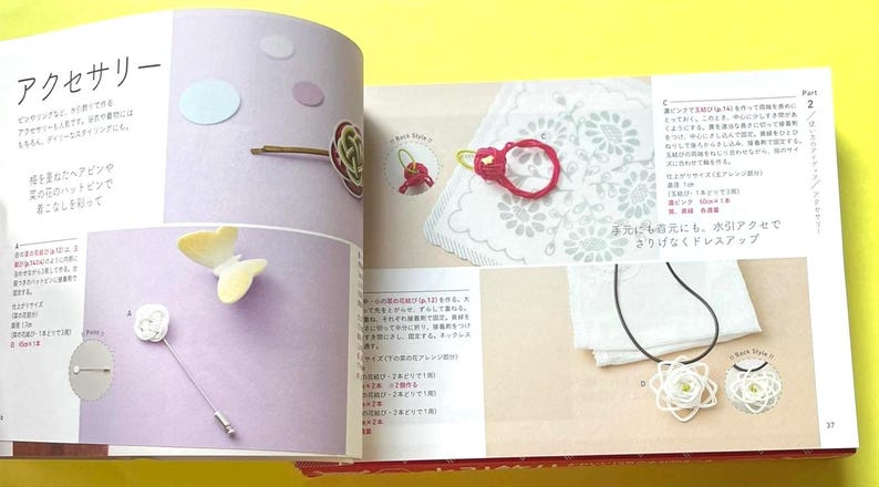 Puede incluir: P&aacute;gina de un libro de manualidades que muestra accesorios hechos a mano. Se incluyen una horquilla, un broche de mariposa, un anillo rojo y un collar con un colgante de flor. La p&aacute;gina tiene texto en japon&eacute;s y est&aacute; sobre un fondo lila claro.