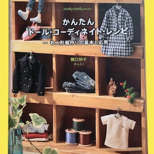 May include: A wooden display unit showcasing miniature doll clothing and accessories. The items include a black jacket, jeans, a plaid shirt, and a white t-shirt. The shelf also holds spools of thread, a doll, and a book titled "Dolly-Dolly Books".