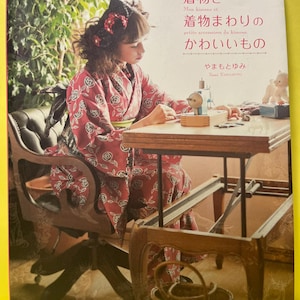 Könnte beinhalten: Buchcover mit einer Frau in einem roten Blumenkimono, die an einem Holztisch sitzt. Der Buchtitel ist auf Japanisch, mit englischen Untertiteln. Eine kleine Nähmaschine und ein Stofftier befinden sich auf dem Tisch.