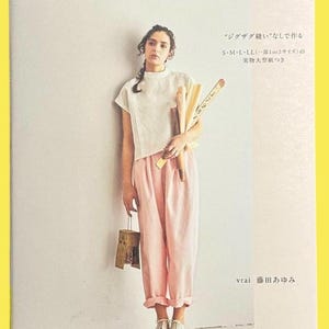 以下が含まれることがあります： 「丁寧に作って長く着る服」というタイトルのソフトカバー本。白い半袖トップス、淡いピンクのパンツ、白いレースアップシューズを着用した人物が写っている。表紙にはS・M・L・LLのサイズ表記や著者名などの日本語テキストが記載されており、木製の道具を持っている。