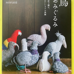 May include: A craft book titled My Favorite Bird Amigurumi by soruni featuring a collection of knitted bird figures including a red and yellow parrot, several grey pigeons, a white bird, and a light pink bird, all displayed on a wooden surface.