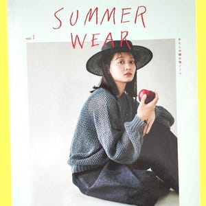 May include: A magazine cover with the words "SUMMER WEAR" in red. A person wearing a black hat, gray sweater, and dark pants holds a red apple. The magazine is labeled "vol. 1" and has Japanese text.