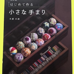 Puede incluir: Un libro japonés titulado "Hacer pequeñas bolas Temari", con bolas decorativas coloridas exhibidas en una caja de madera. Las bolas presentan patrones intrincados en varios colores, incluyendo rojo, azul y verde. El libro forma parte de la "Lady Boutique Series".