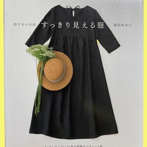 以下が含まれることがあります： 黒いワンピース、丸いネックラインと七分袖。本の上に表示されています。麦わら帽子と緑の植物がワンピースの前に置かれています。表紙には日本語のテキストが表示されています。