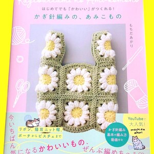 May include: A crochet book cover featuring a daisy-patterned tote bag. The bag is made of green and white yarn with yellow centers. The book title is in Japanese, with additional text and illustrations of yarn, scissors, and a cat.