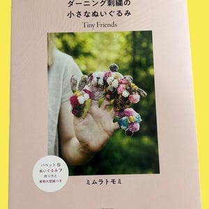 Può includere: Copertina di un libro con il titolo "Tiny Friends" in inglese e giapponese. La copertina presenta una foto di animali di peluche ricamati in miniatura e colorati. Il libro include anche le istruzioni per realizzare i giocattoli.