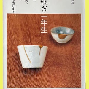 May include: A book cover featuring Japanese text and two ceramic bowls. The text includes "Kintsugi First Year Student". The bowls are white and blue, with gold detailing, showcasing the art of Kintsugi.