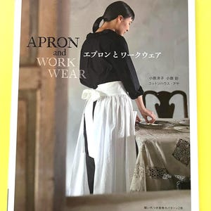 以下が含まれることがあります： 白いエプロンと黒いトップスを着た女性が写っている本の表紙。「APRON and WORK WEAR」というタイトルが英語で書かれており、日本語のテキストも含まれています。背景はニュートラルな色調のインテリアです。