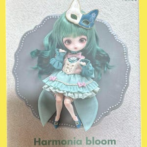 May include: A magazine cover featuring a doll with green hair and a blue and white mask. The doll is wearing a blue and pink dress. The magazine title is "Dollybird" with the text "vol.33". The text "Harmonia bloom" and "KUMAKO" are also visible.