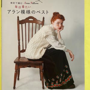 以下が含まれることがあります： applemintsと題された日本語と英語のテキストが記載された手芸本。表紙には、白いシャツの上にクリーム色のケーブルニットベストを着用し、赤い花柄の刺繍が施された長い黒のスカートを履いた人物が木製の椅子に座っている様子が写っています。