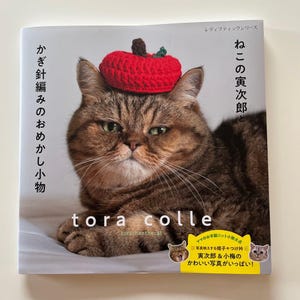 以下が含まれることがあります： 赤いかぎ針編みのリンゴの帽子をかぶった茶色のトラ猫の表紙。 書名「tora colle」と上下に日本語のテキスト。 追加のテキストとイラストがあります。