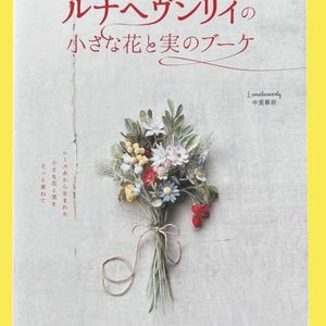 Può includere: Copertina di un libro con un bouquet all'uncinetto di piccoli fiori e bacche. Il titolo è in giapponese. Il bouquet comprende fiori bianchi, rossi, gialli e blu, legato con un nastro. Il nome dell'autore è visibile.