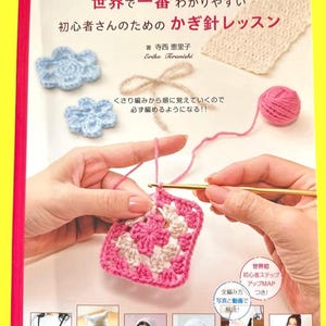 以下が含まれることがあります： 日本のテキストと編み物の写真が掲載された本の表紙。表紙には、ピンクと白のかぎ針編みの正方形、青いかぎ針編みの花、ピンクの糸玉が描かれています。タイトルは「初心者向けのかぎ針編みレッスン」です。