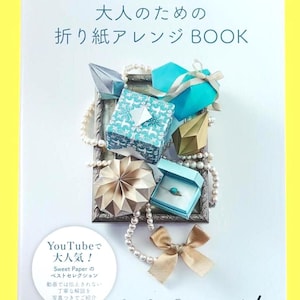 Może przedstawiać: Okładka książki z japońskim tekstem i sztuką origami. Tytuł brzmi "Książka aranżacji origami dla dorosłych". Prezentowane są modele origami w różnych kształtach i kolorach, w tym niebieskie, złote i wzorzyste pudełka.