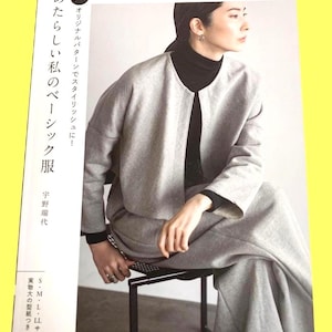 以下が含まれることがあります： 黒いタートルネックを着て、ライトグレーのジャケットとパンツを着た女性が、黒い金属製の椅子に座っています。 ジャケットは前開きです。 画像は日本のファッション雑誌からのもので、日本語のテキストが表示されています。