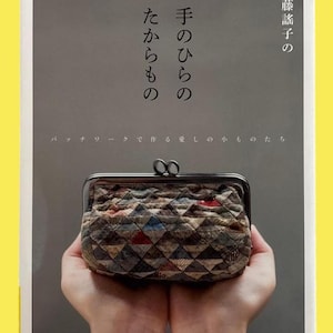 以下が含まれることがあります： 手作りのパッチワークの小銭入れが手に持たれた本の表紙。小銭入れは茶色、青、赤の色合いの幾何学模様。表紙には日本語のテキストが書かれています。