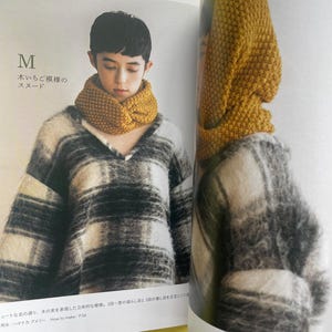 May include: A mustard yellow knitted snood with a textured, bumpy stitch pattern worn around the neck. The snood is featured in a book page alongside a black and white plaid, fuzzy, long-sleeved sweater. The text M and Japanese characters are visible.