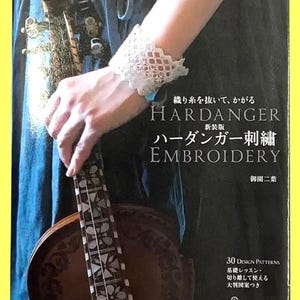 以下が含まれることがあります： 日本語で「HARDANGER EMBROIDERY」というタイトルの本。白いレースのカフスと弦楽器が特徴です。表紙には英語と日本語のテキストがあります。画像には、楽器のネックの詳細なビューも含まれています。