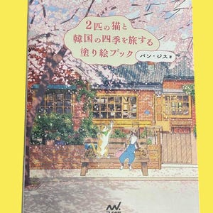 Op de afbeelding: Een Japans kleurboek met een omslagillustratie van twee katten op een bank voor een gebouw, met kersenbloesem op de achtergrond. De titel is in het Japans.