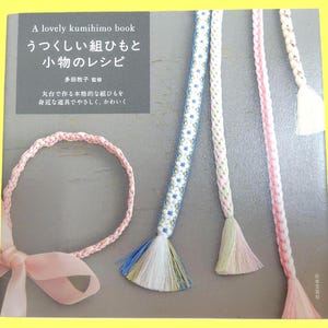 May include: A book titled "A lovely kumihimo book" with Japanese text and examples of braided cords in pink, white, and blue. The cords have decorative tassels. A pink braided cord with a bow is also shown.