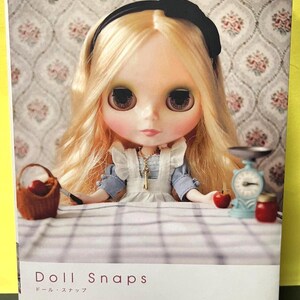 Puede incluir: La portada del libro "Doll Snaps" presenta una muñeca con cabello rubio, ojos grandes y una diadema negra. La muñeca lleva un vestido azul y blanco. El fondo tiene un estampado floral.
