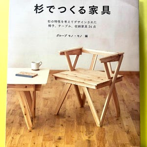 May include: A Japanese book titled "Furniture Made of Cedar", showcasing a wooden chair and table. The cover features Japanese text and furniture illustrations. A mug and coaster are on the table. The book is on a wooden floor.